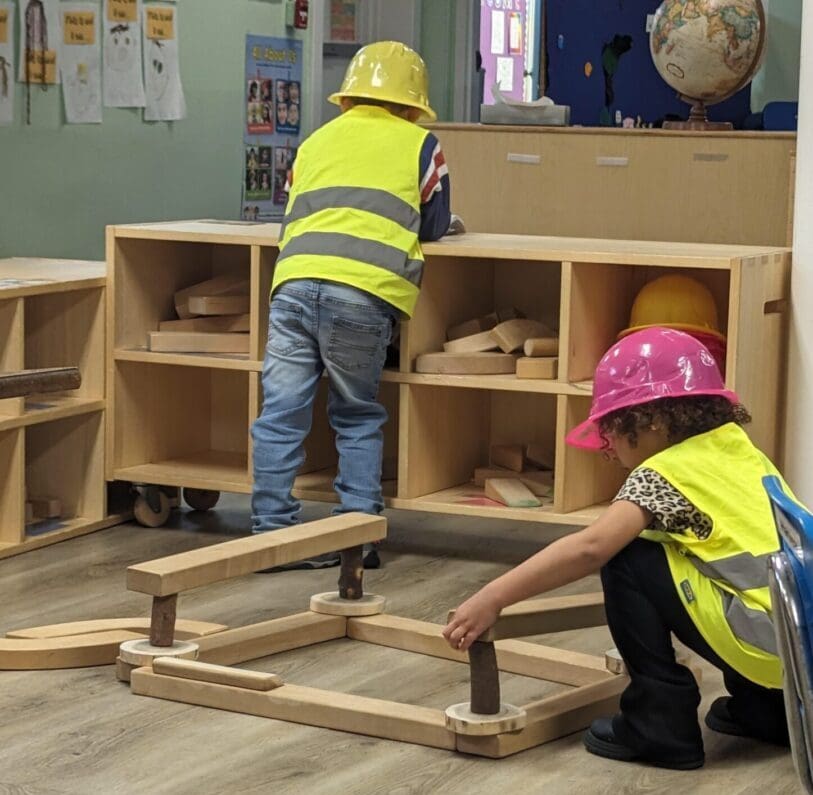 Two children in yellow vests and hard hats work on a wooden shelf.